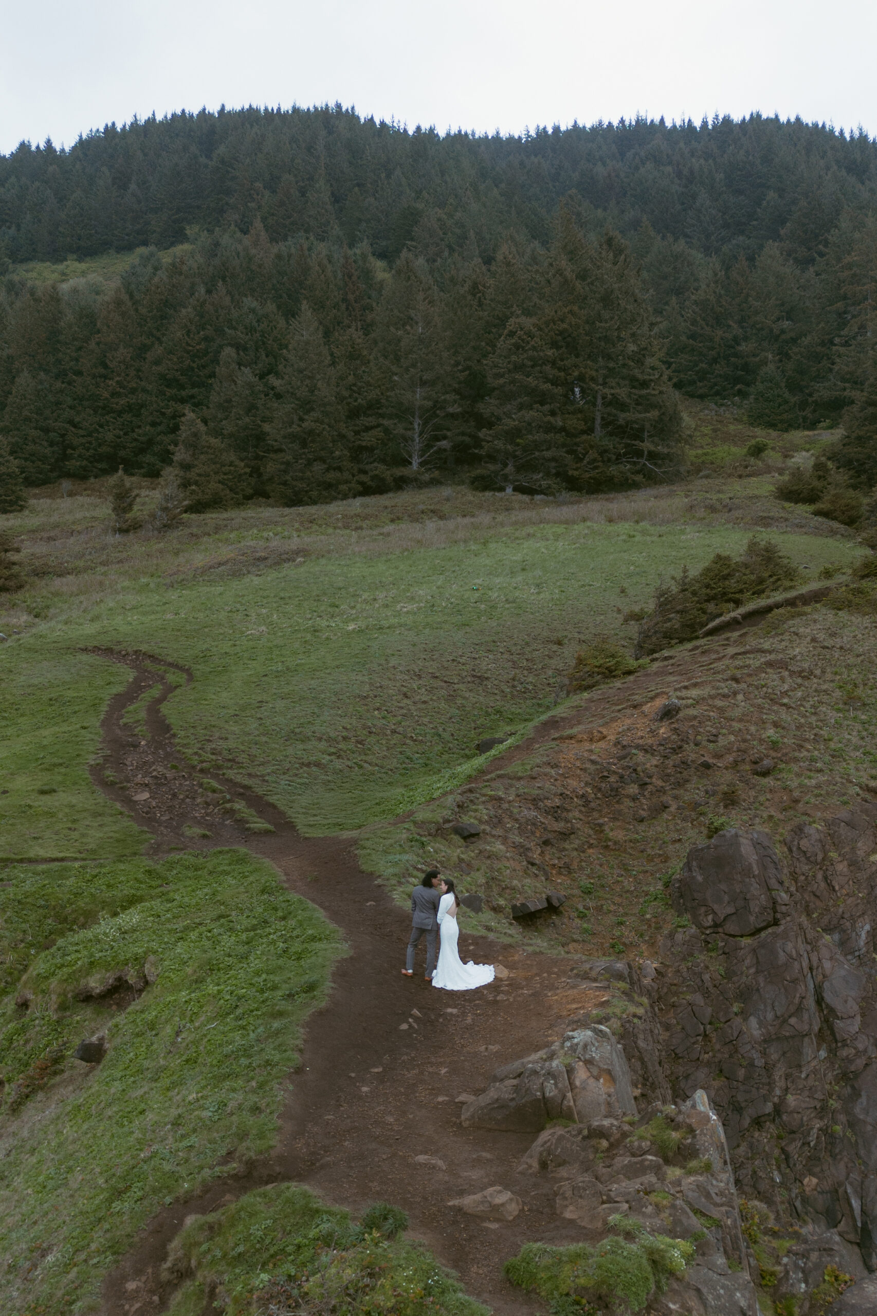 A couple embraces on a winding trail surrounded by lush green hills and dense forest, with the bride in white and the groom in a suit.