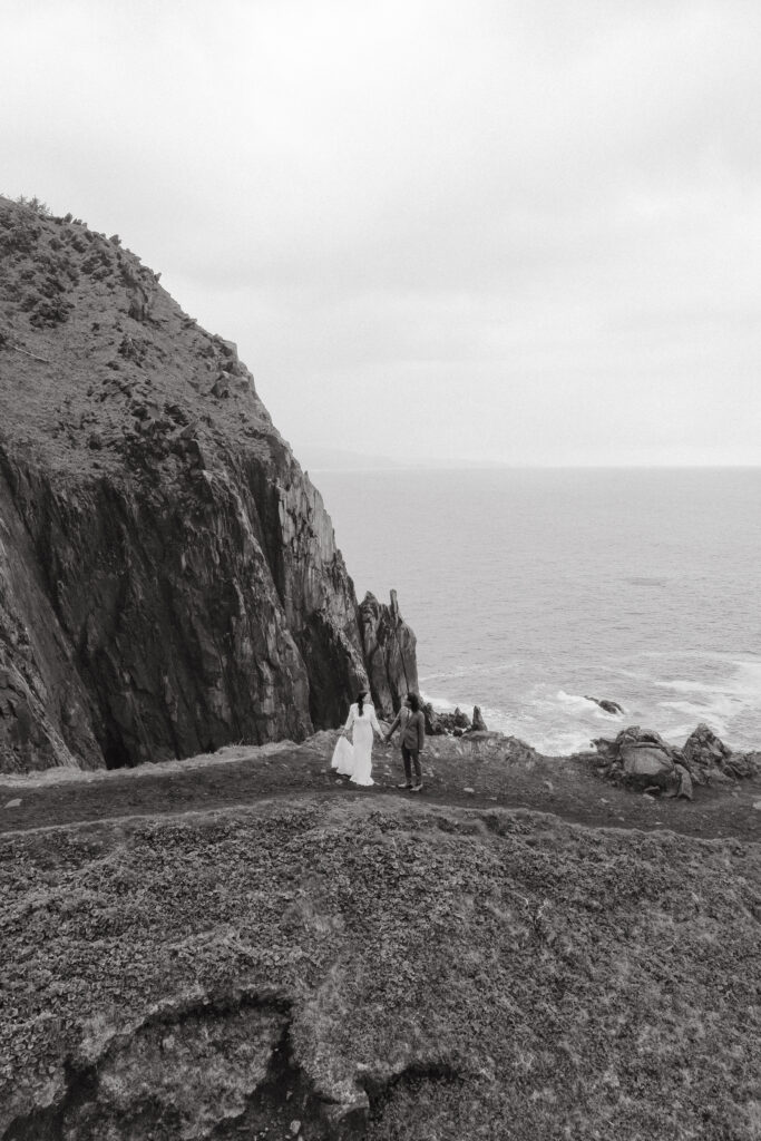 A couple stands hand in hand near the edge of a rugged cliffside, looking out at the ocean beneath a cloudy sky.