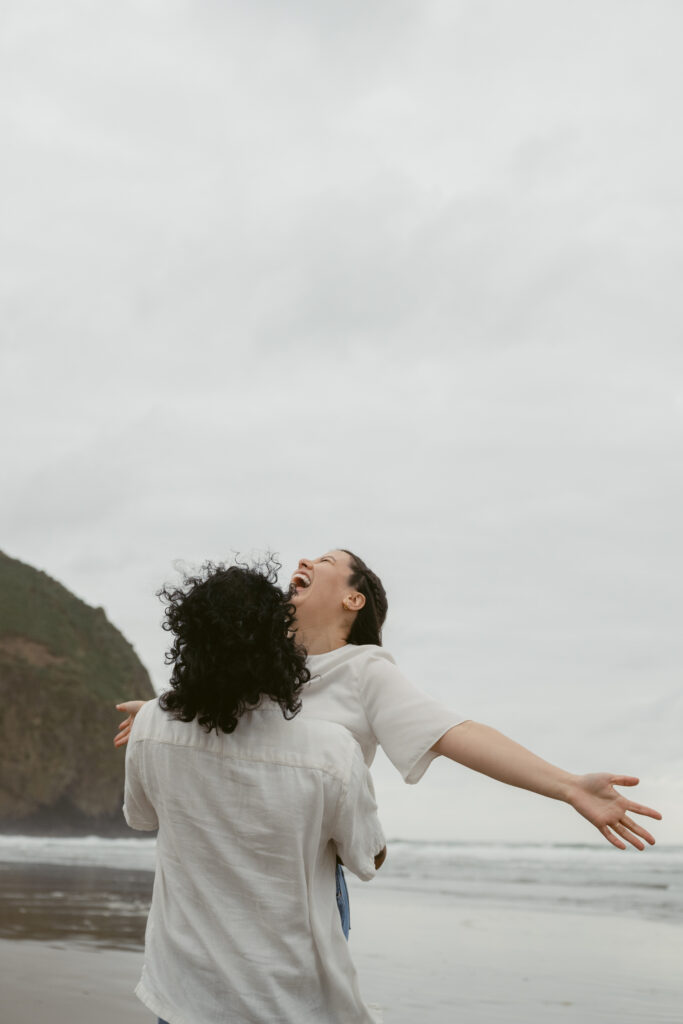 Couple laughing and spinning on the beach during their Oregon Coast elopement, with ocean in background.