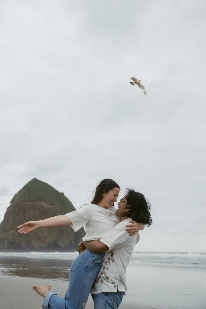 Couple laughing and lifting each other during their Oregon Coast elopement, with Haystack Rock behind them.