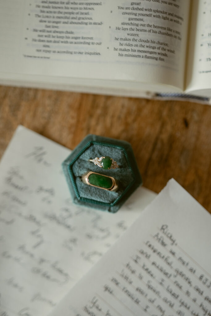 Green wedding rings in a velvet box, surrounded by handwritten vows and an open Bible.