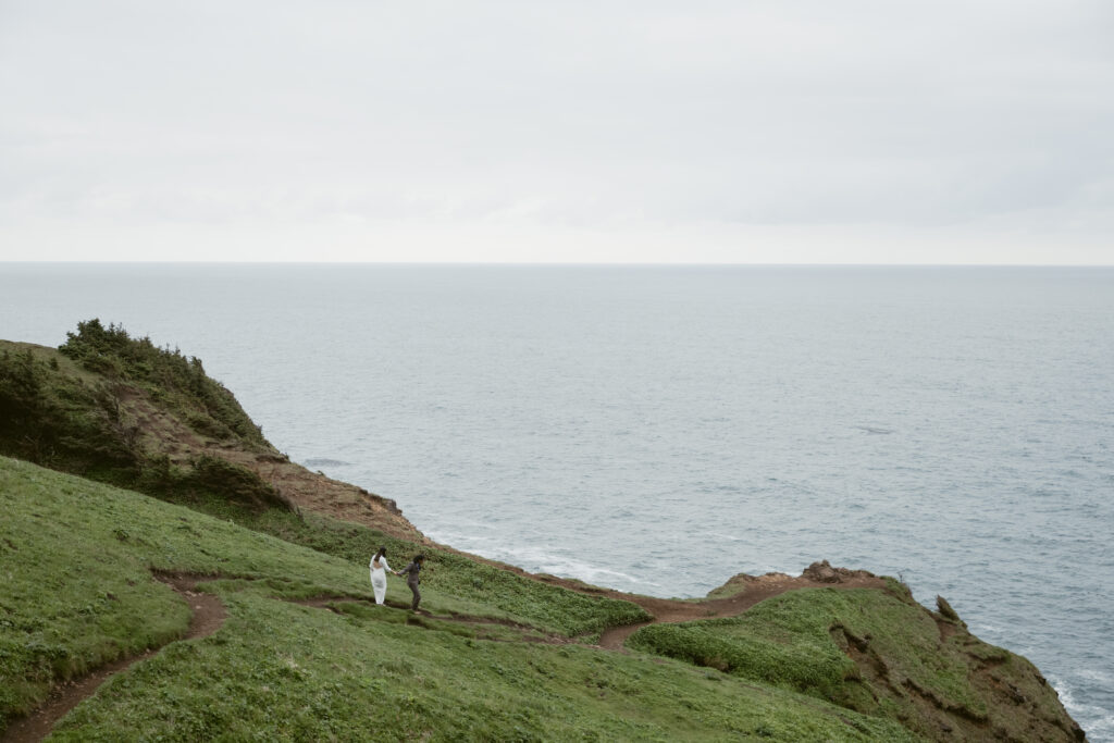 A couple walks hand-in-hand along a winding trail on a cliffside, overlooking the vast and serene ocean. The overcast sky and lush green landscape create a peaceful, intimate atmosphere, capturing a quiet moment of connection in nature.