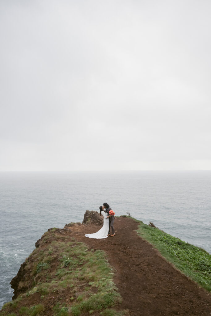 A couple shares a kiss on a cliffside trail overlooking the ocean, with the bride in a flowing white gown and the groom holding a bouquet.