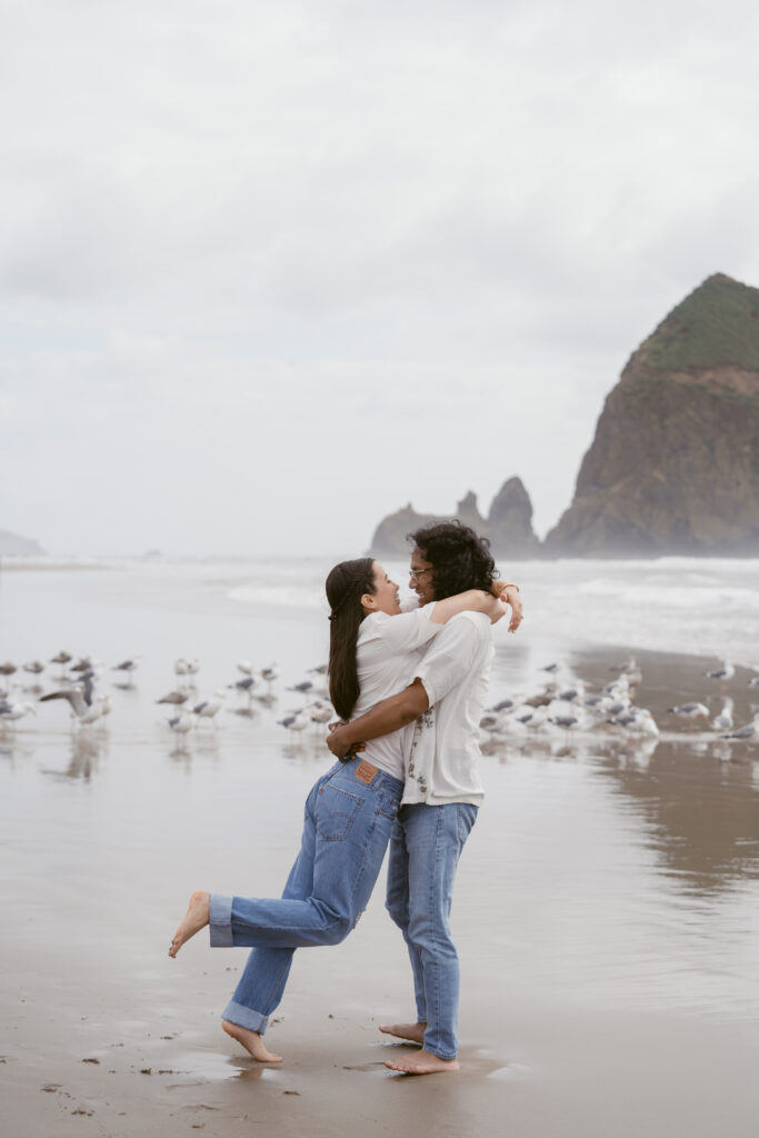 Couple laughing and dancing barefoot during their Oregon Coast elopement, surrounded by seagulls on the beach.