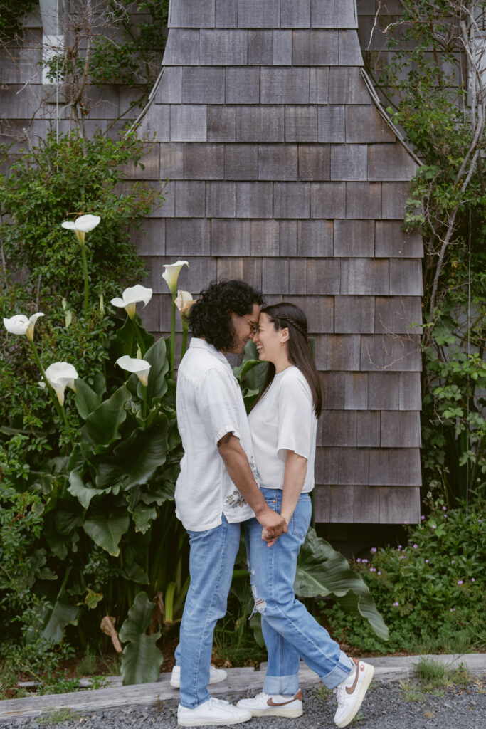 Couple standing forehead to forehead, holding hands and smiling in front of a shingled wall and flowers.