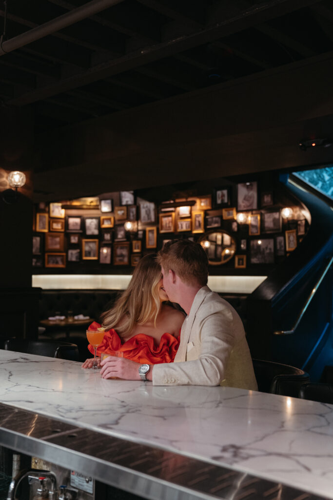 engagement-photo-tips Couple sitting at a marble bar, sharing an intimate moment while enjoying cocktails, with a warm, moody gallery wall in the background.