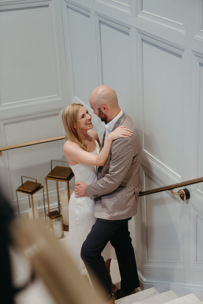 engagement-photo-tips Couple embracing and smiling at each other on a staircase with white paneled walls and modern gold lanterns, sharing a romantic moment.