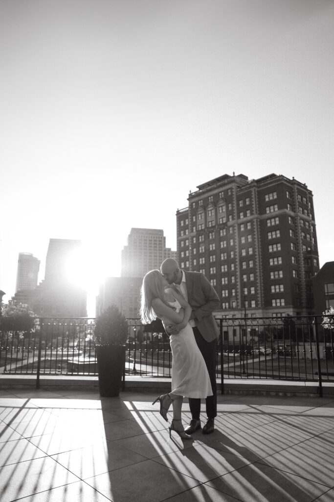 engagement-photo-tips Couple kissing on a rooftop at sunset, with dramatic shadows and Cincinnati city buildings in the background, captured in black and white.
