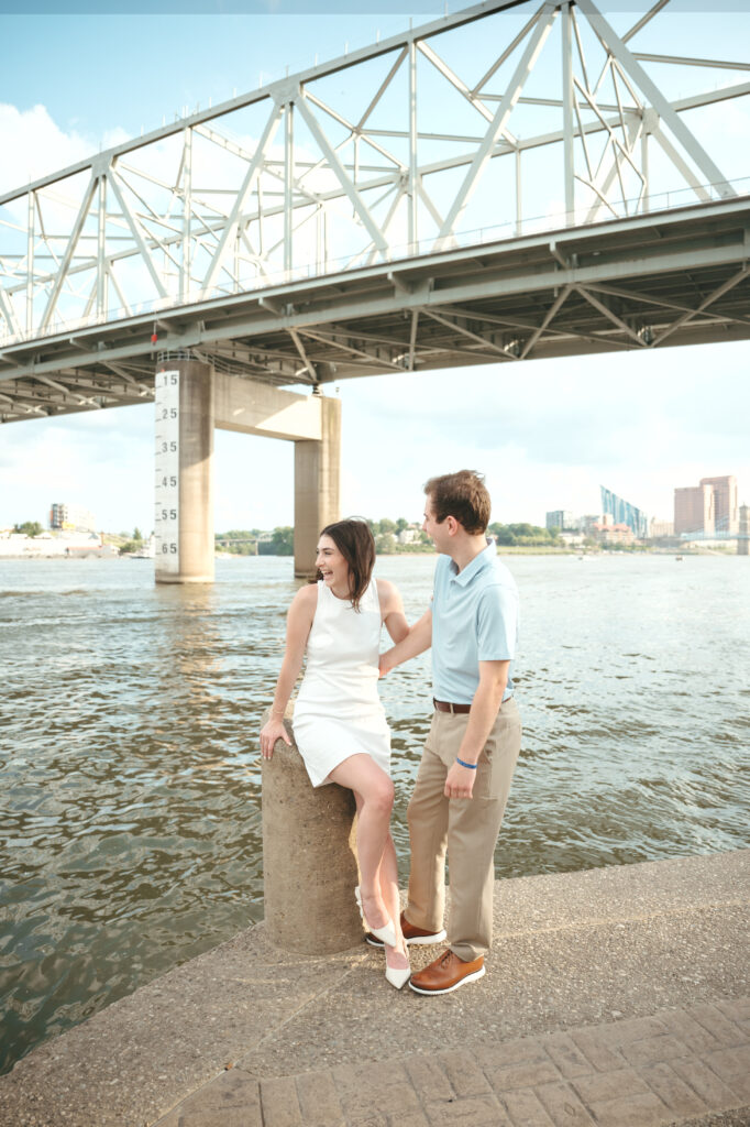 engagement-photo-tips Couple enjoying a lighthearted moment by the riverfront during their Cincinnati engagement session near the bridge, captured as part of creative engagement photo tips.