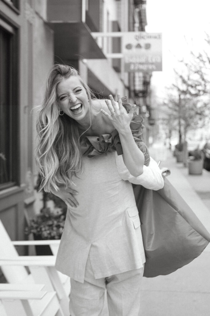 engagement-photo-tips Woman laughing and showing off her engagement ring while being lifted by her partner on a city sidewalk, captured in a joyful candid moment.
