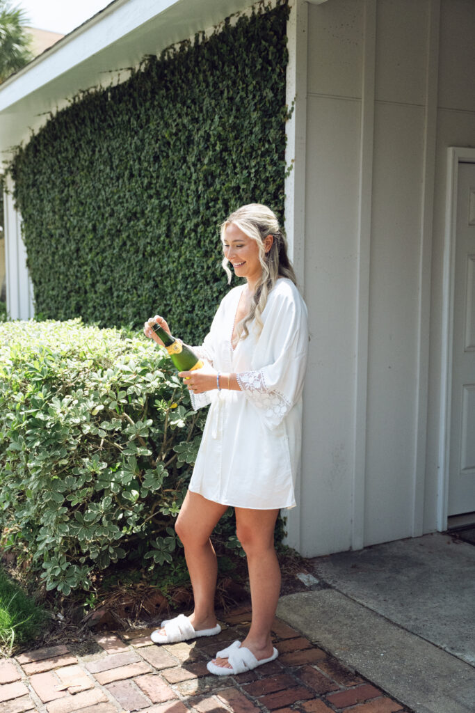 Bride in a white robe and slippers opening champagne outside on the morning of her Georgia wedding, smiling in the sunshine near ivy-covered walls.