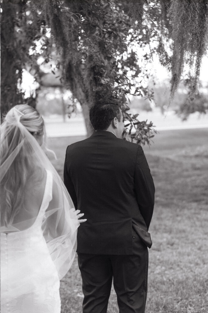 Bride walking up behind the groom for their first look, captured outdoors beneath trees draped in Spanish moss.
