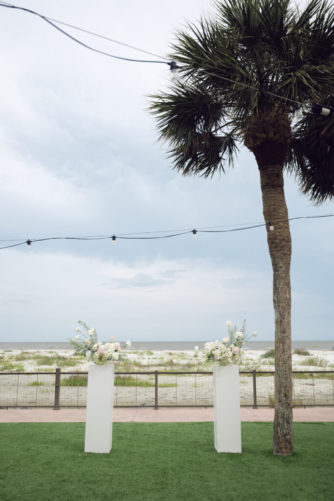 A serene beach wedding ceremony setup with two white floral arrangements on tall pedestals, set against the backdrop of the ocean and a cloudy sky, with string lights and a palm tree adding a coastal charm.
