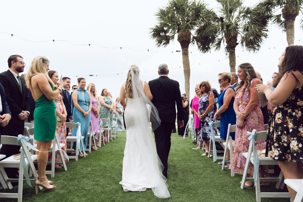 A smiling bride walks arm-in-arm with her father down the aisle, both dressed in formal wedding attire. The bride wears a white gown with a long veil, while her father wears a classic black tuxedo.