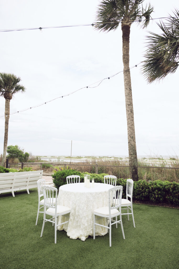Round cocktail table with white lace tablecloth and chairs set under palm trees with string lights, overlooking the beach and ocean.