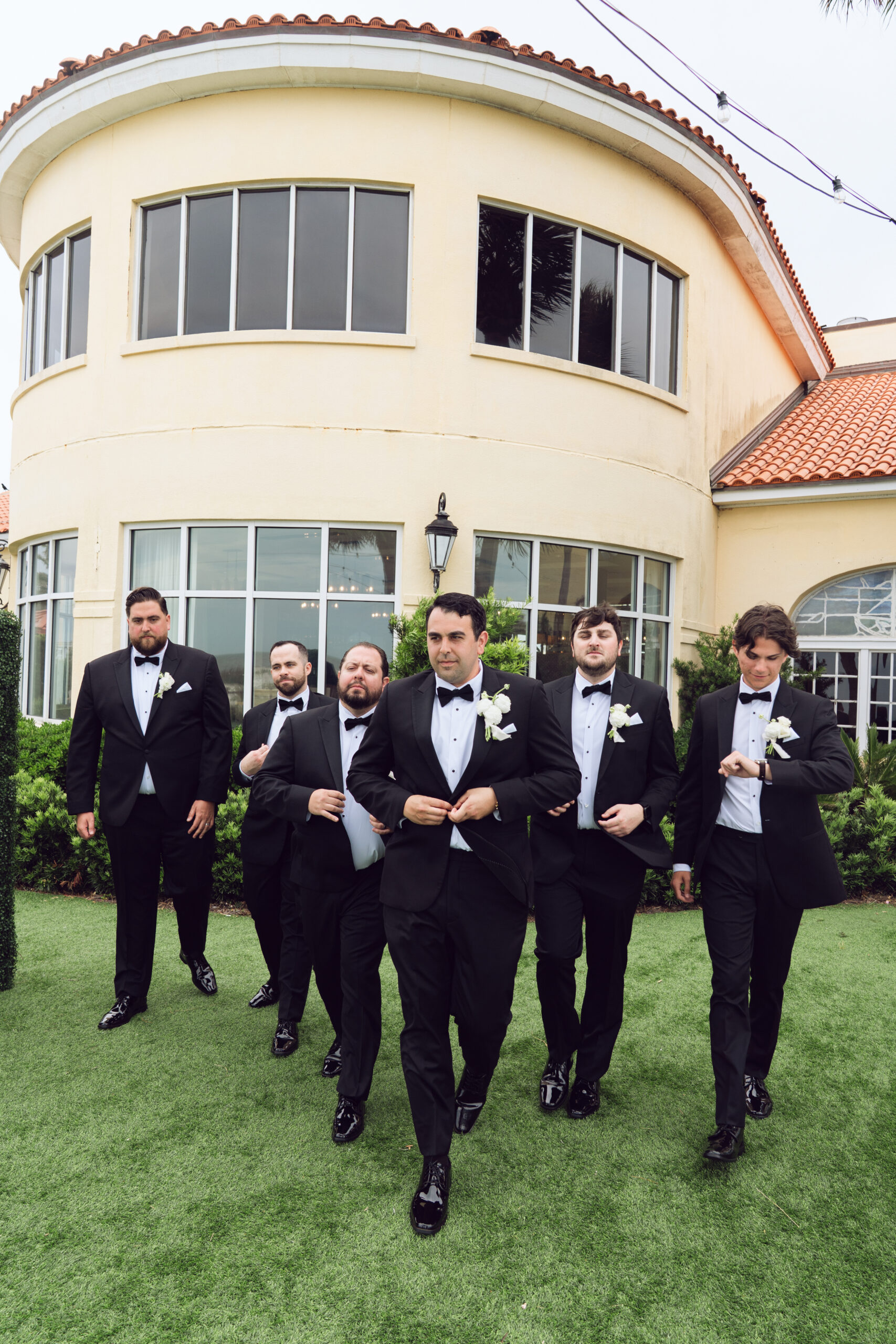 Groom and groomsmen walking confidently in black tuxedos outside a Mediterranean-style venue.