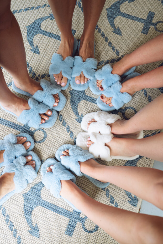 Bridesmaids wearing matching fluffy blue slippers and the bride in white slippers, standing in a circle on an anchor-patterned rug before a Georgia wedding.