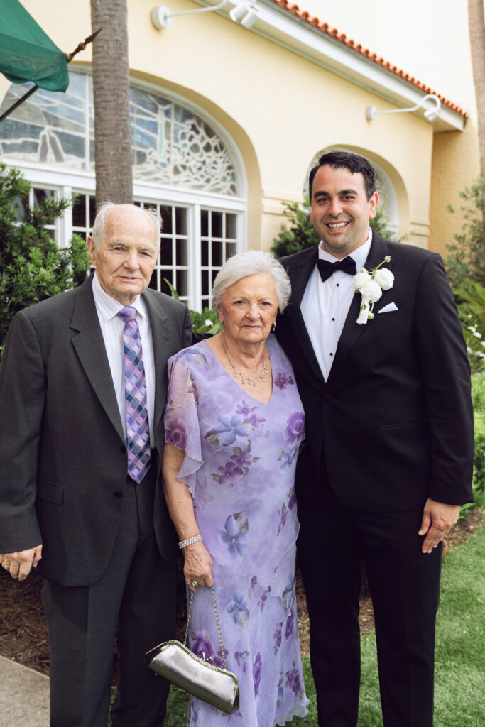 Groom smiling with grandparents, standing outside the venue with greenery and windows behind them.