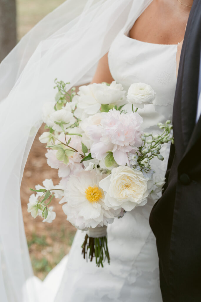 Close-up of the bride’s elegant white bouquet featuring peonies, ranunculus, and delicate greenery, held against her lace gown.