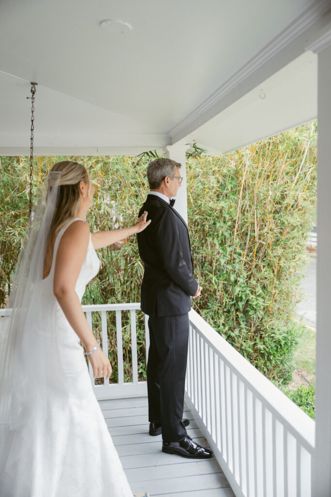 Bride reaches out to tap her father on the shoulder during an emotional first look moment on a cottage porch, surrounded by lush greenery on the morning of her wedding.