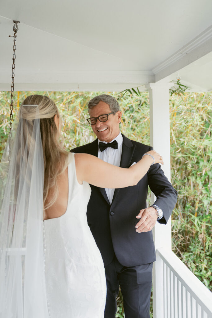 Bride and her father smile as they embrace during a joyful first look on the porch, with greenery in the background on wedding morning.