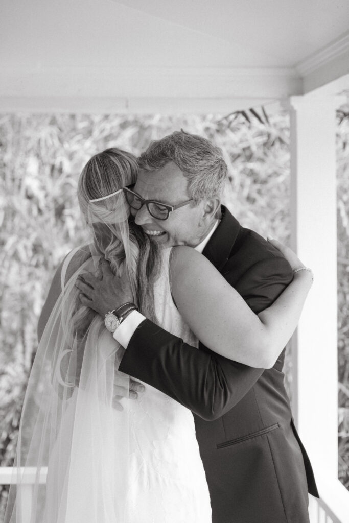Father of the bride hugs his daughter tightly during an emotional first look on the porch, both smiling in a heartfelt black and white moment.
