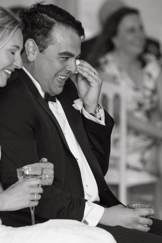 Groom wipes away a tear while laughing during a heartfelt wedding reception toast.