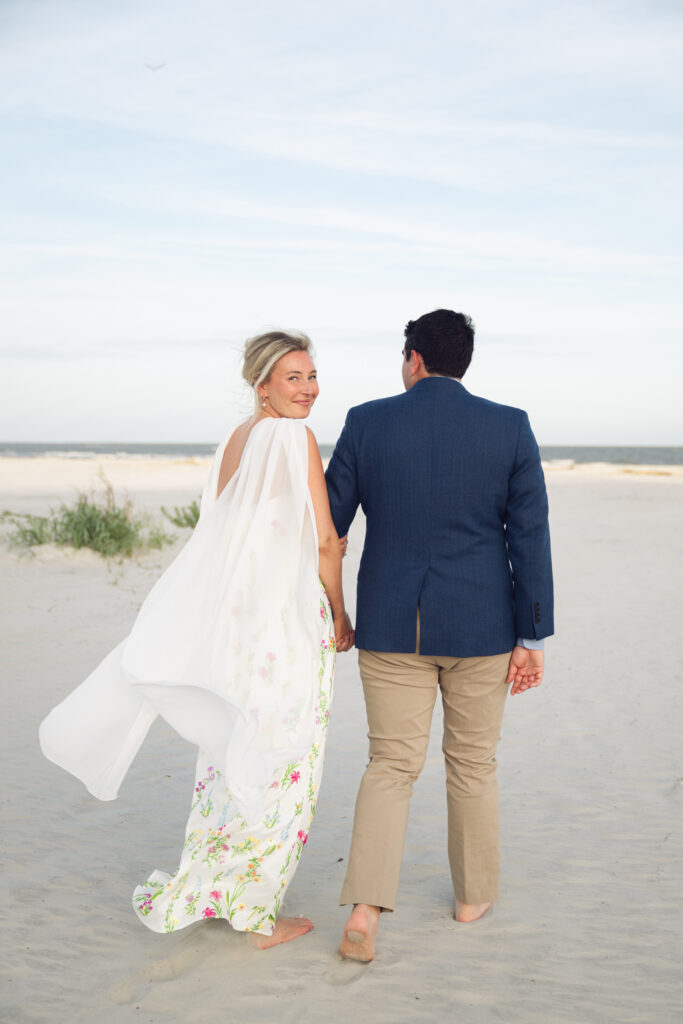 Bride and groom walking barefoot on the beach at sunset during their Georgia wedding, with the bride smiling back and her dress flowing in the breeze.