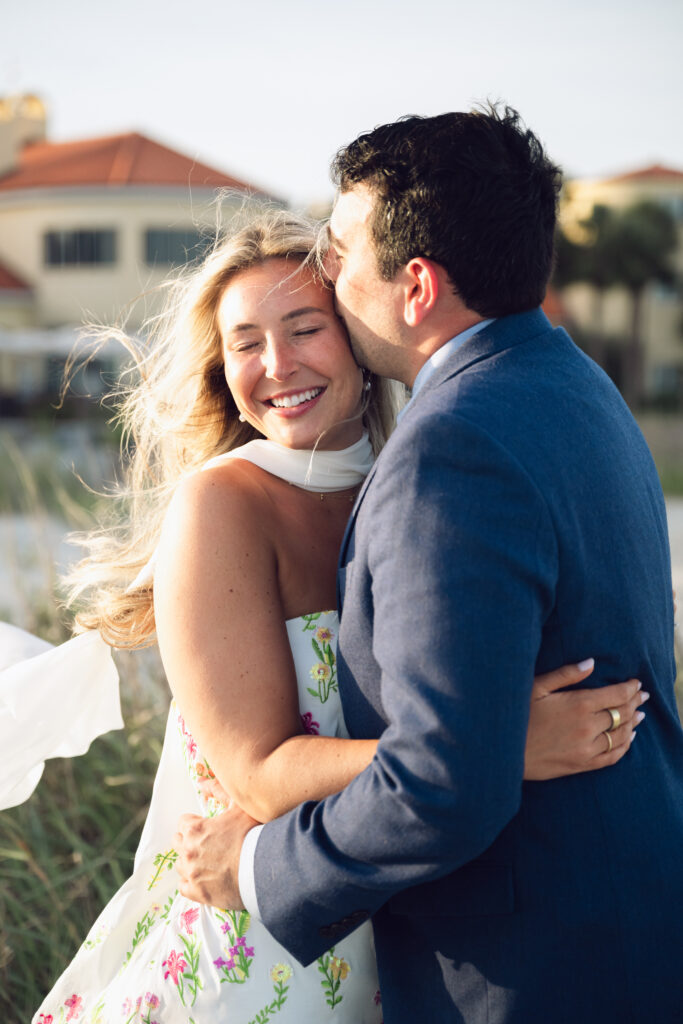 Groom kisses bride on the forehead as she smiles with eyes closed, wrapped in sunlight during their joyful Georgia wedding by the coast.