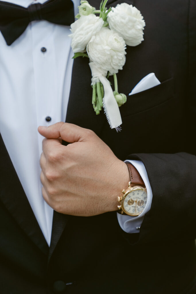 Close-up of the groom’s hand adjusting his black tuxedo jacket, showing a gold watch and white boutonniere made of ranunculus flowers.