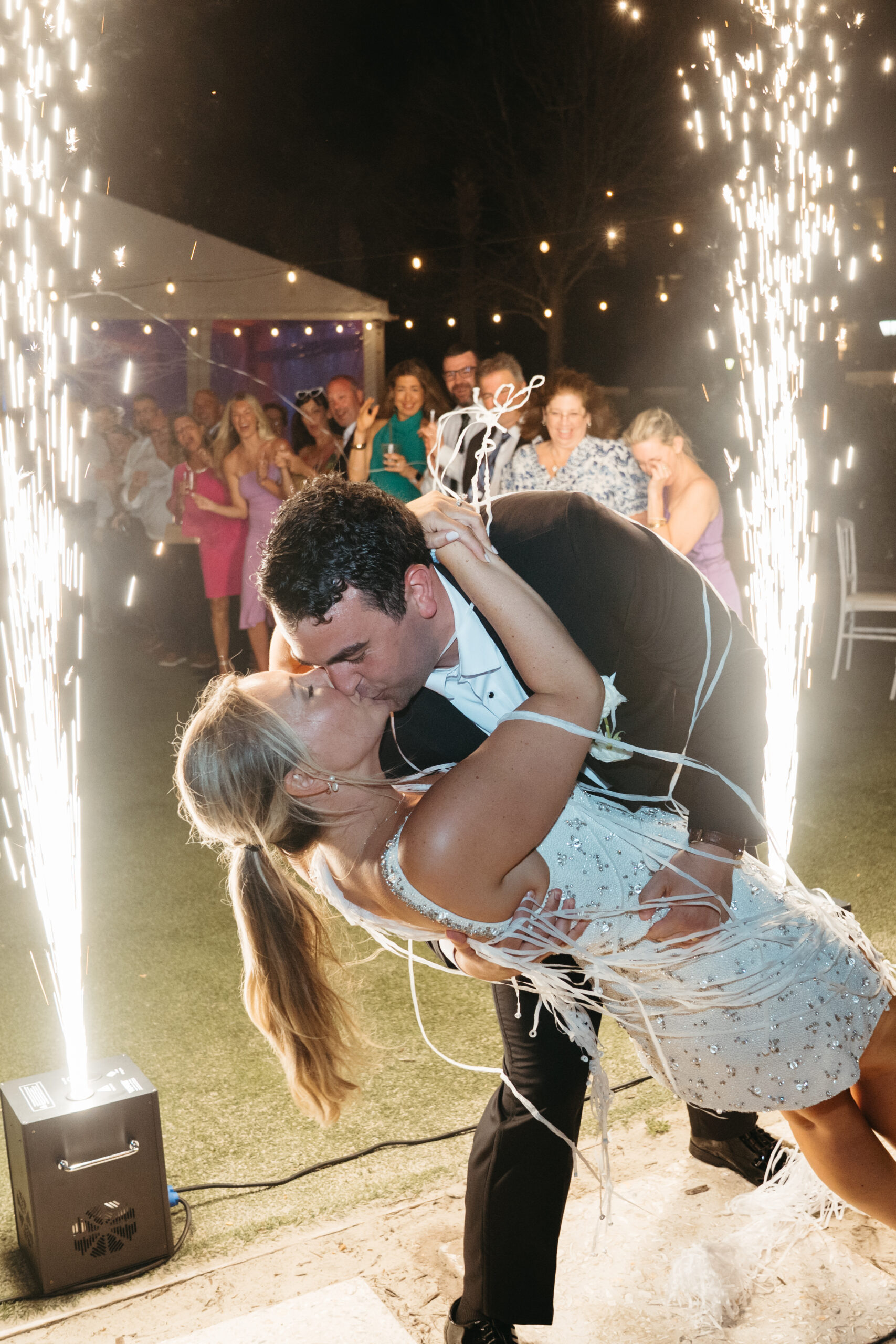 Newlyweds share a dramatic dip kiss during their grand exit with cold sparks and cheering guests.