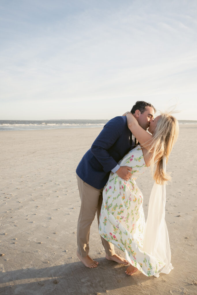 Groom dips and kisses bride on the beach during their Georgia wedding, with sunlight catching her flowing floral dress and scarf in the breeze.