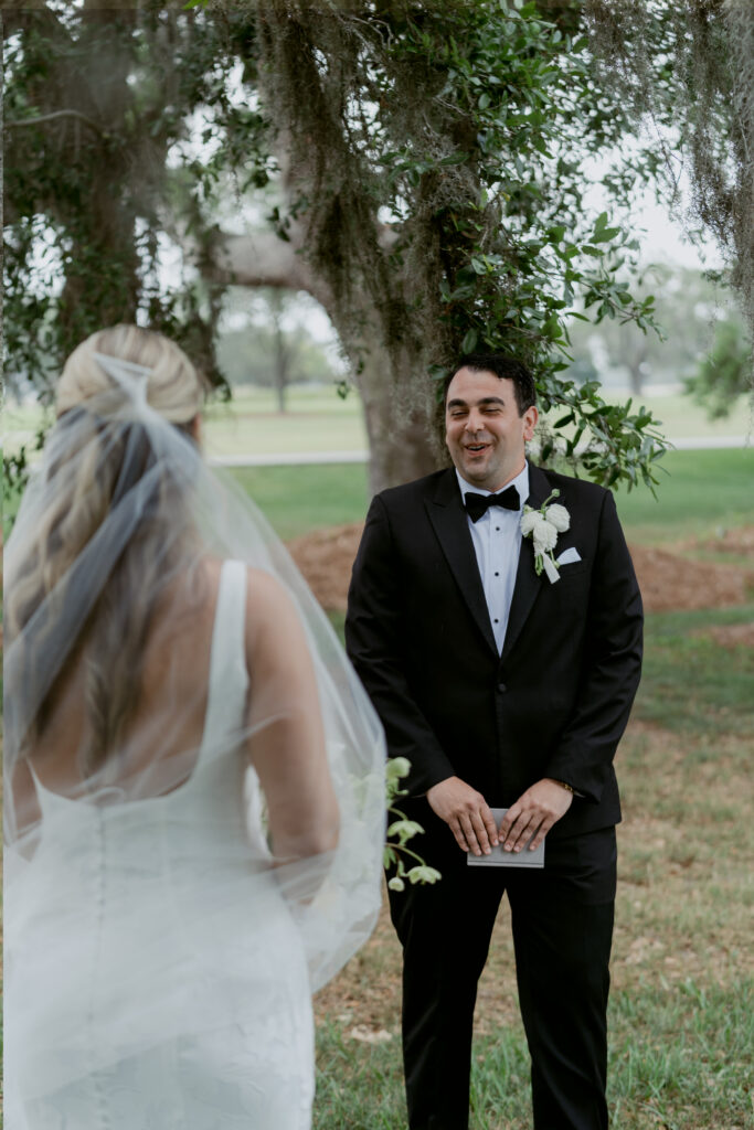 Groom smiling emotionally as he sees his bride for the first time during their outdoor first look under trees with hanging Spanish moss.