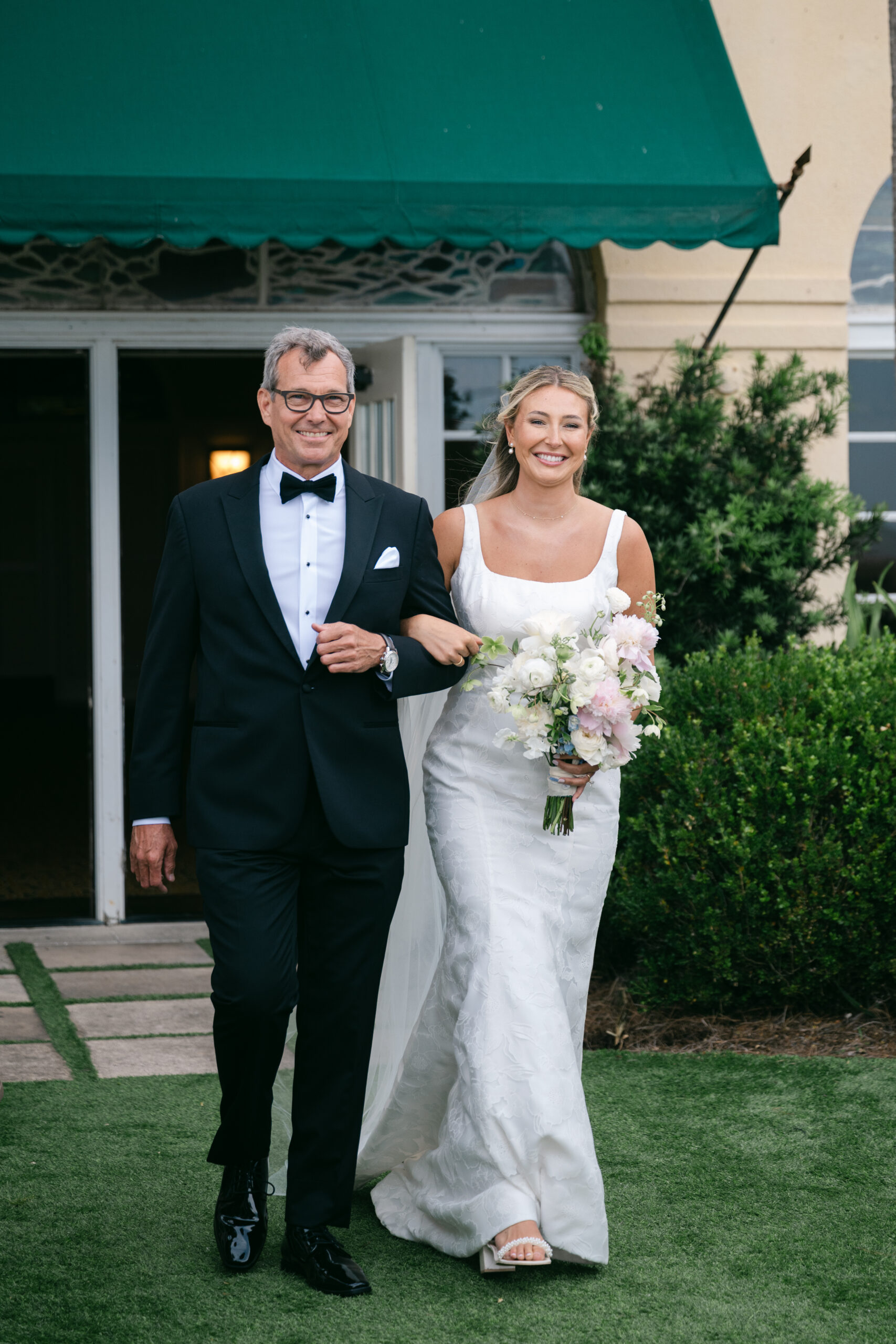 A smiling bride walks arm-in-arm with her father down the aisle, both dressed in formal wedding attire. The bride holds a pastel bouquet and wears a white gown with a long veil, while her father wears a classic black tuxedo.