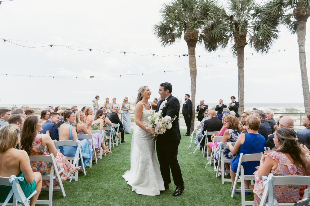 Bride and groom walk up the aisle smiling just after their beachside ceremony, surrounded by guests seated on either side under string lights and palm trees.