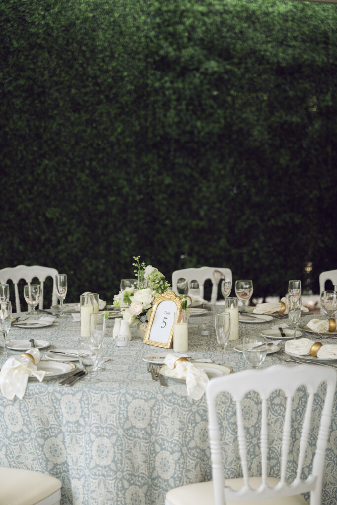 Close-up of a reception table with floral centerpiece, candles, and a gold-framed table number five, surrounded by white chairs and patterned linens.
