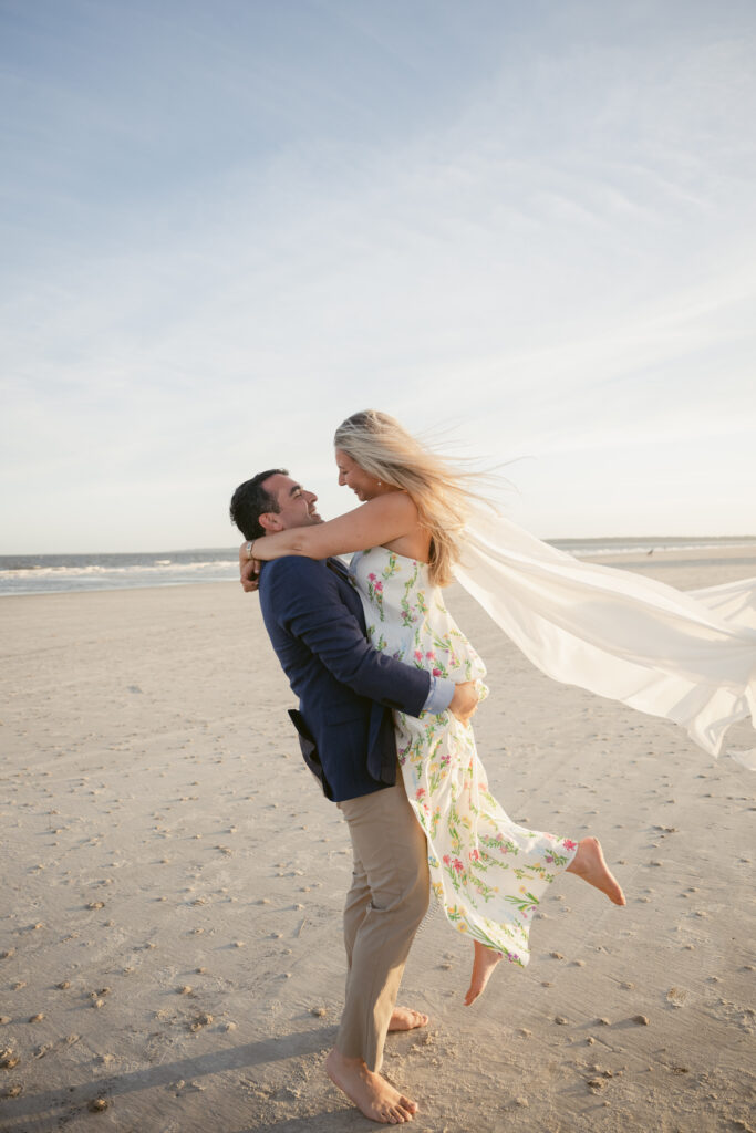 Groom lifts bride on the beach during their Georgia wedding, with her floral dress and scarf flowing in the ocean breeze under golden sunlight.