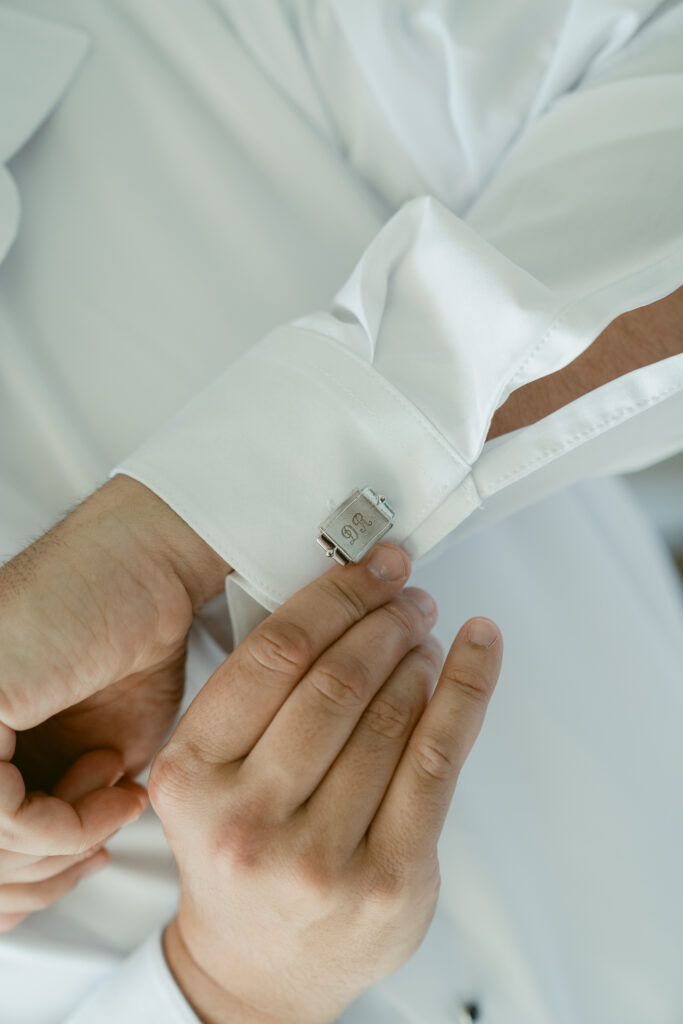 Close-up of the groom fastening personalized silver cufflinks with engraved initials and wedding date on his white dress shirt.