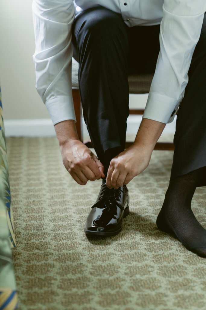 Groom tying the laces of his black patent leather dress shoes while sitting on a chair, dressed in a white shirt and black pants.