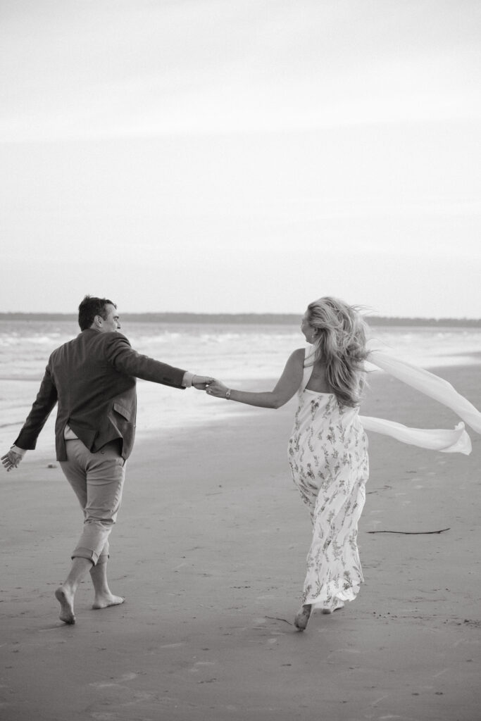 Black and white image of a couple running barefoot along the shoreline, holding hands during their joyful Georgia wedding beach session.