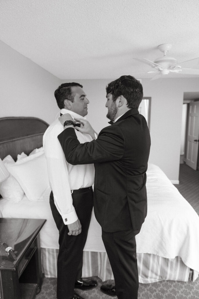 Groomsman helps the groom adjust his bow tie in a hotel room as they get ready for the wedding, both wearing formal attire.