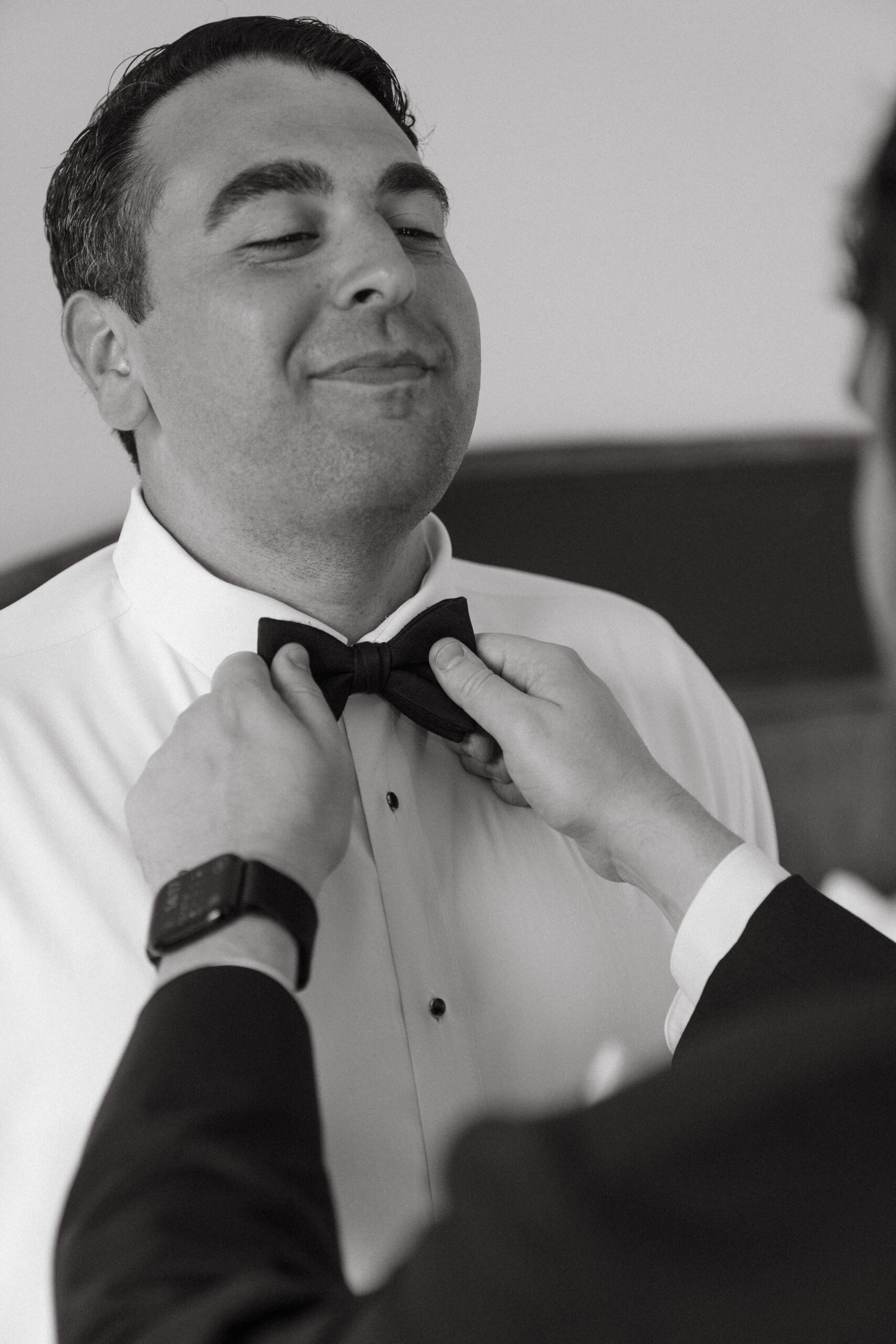 Groom smiles proudly while a groomsman helps adjust his bow tie, captured in a candid black and white getting ready moment.