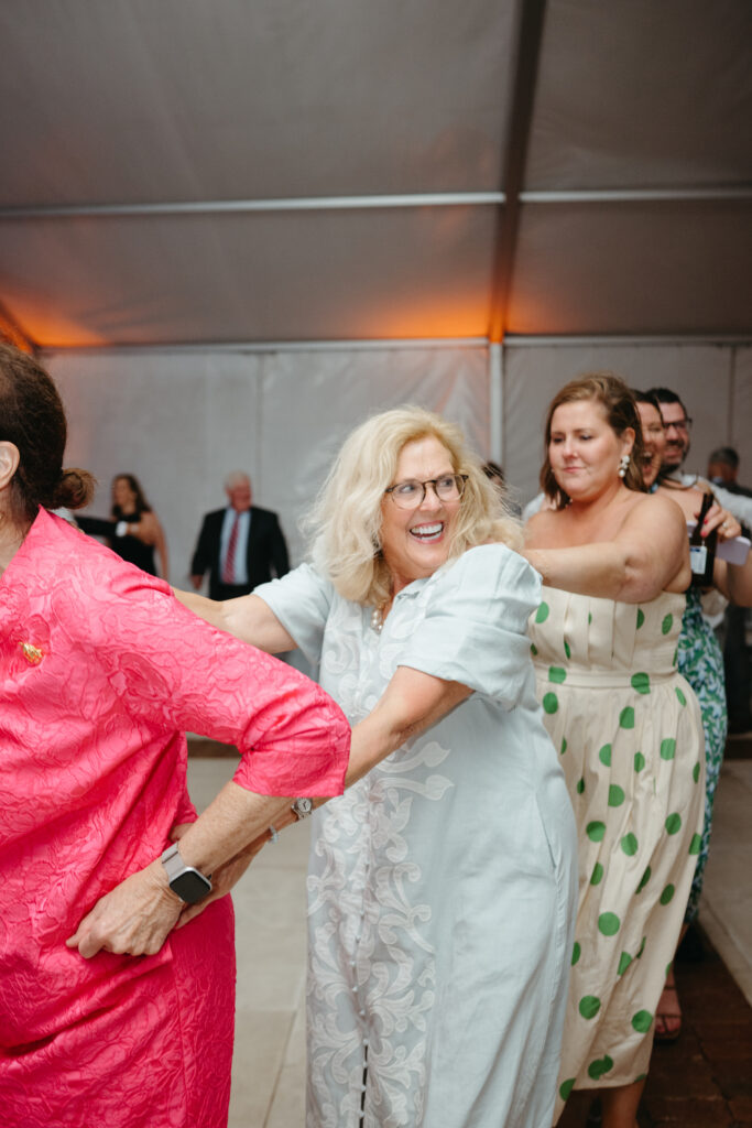 Guests laugh and dance in a conga line during the wedding reception under a tent.