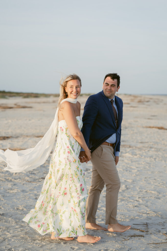 Bride and groom walk hand in hand along the beach during their Georgia wedding, with the bride smiling over her shoulder in a floral dress.