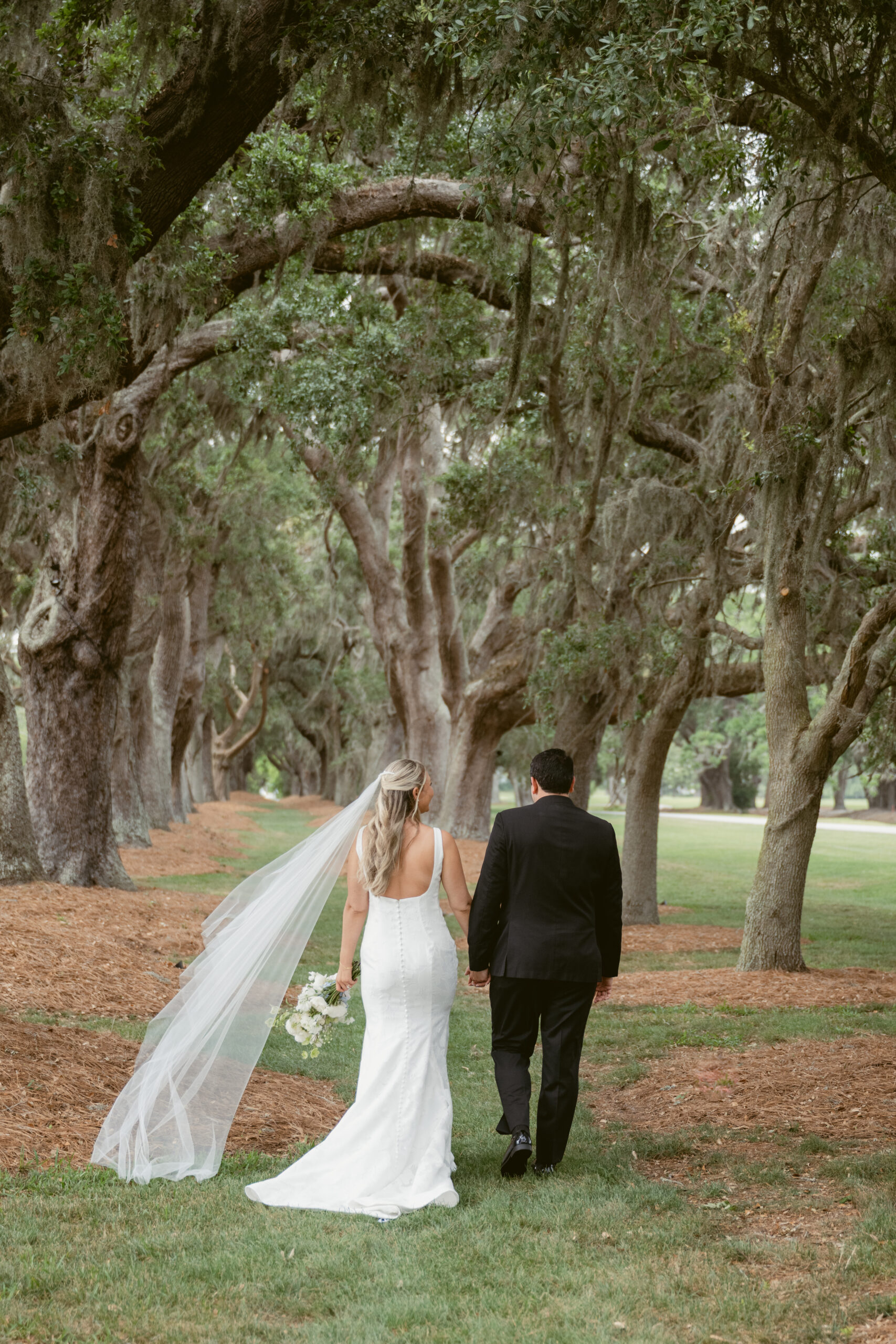 Bride and groom walk hand in hand down a tree-lined path, with the bride’s veil trailing behind and creating a soft, romantic flow.