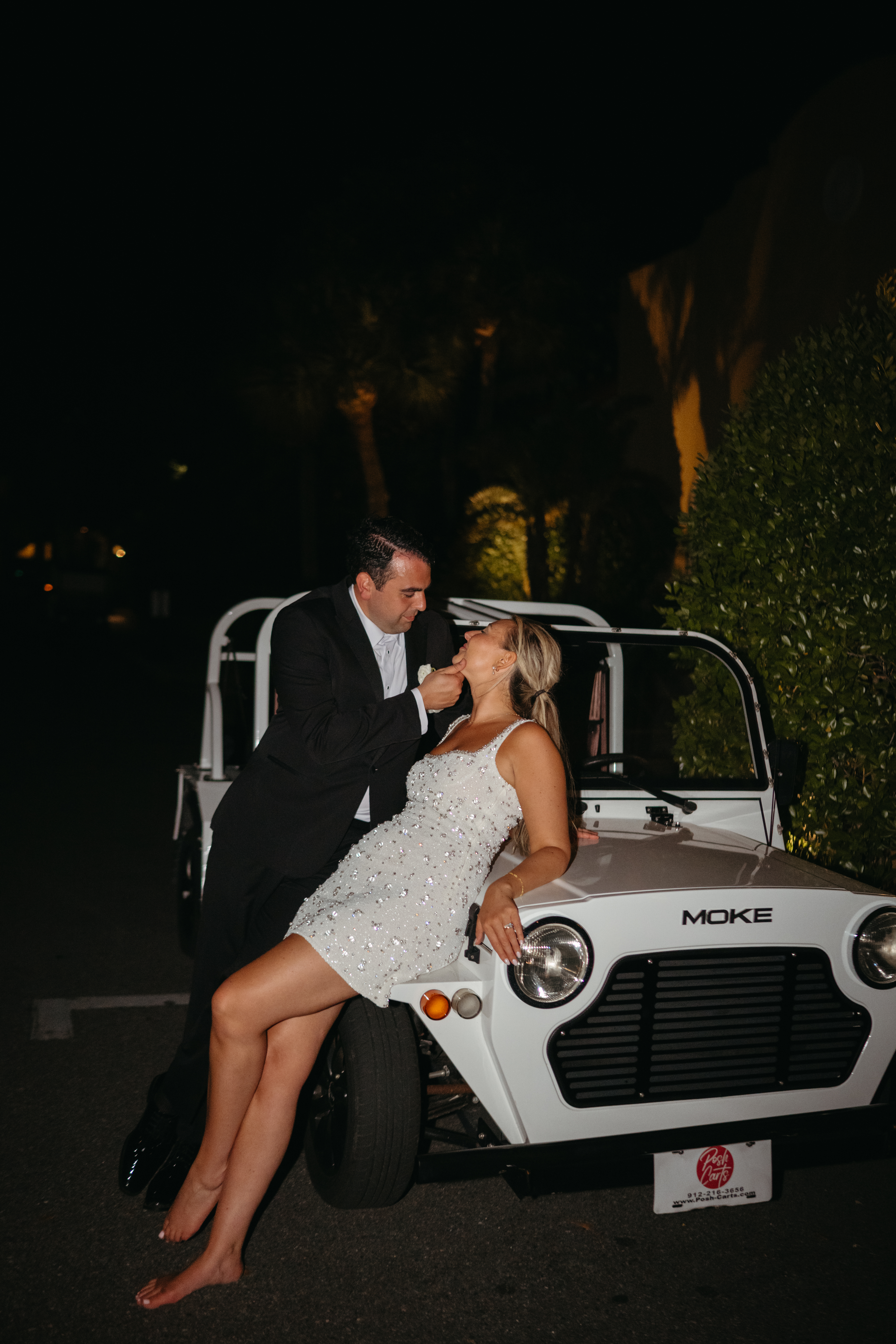 Bride in sparkly dress leans on white Moke car while groom tenderly cups her chin at night.