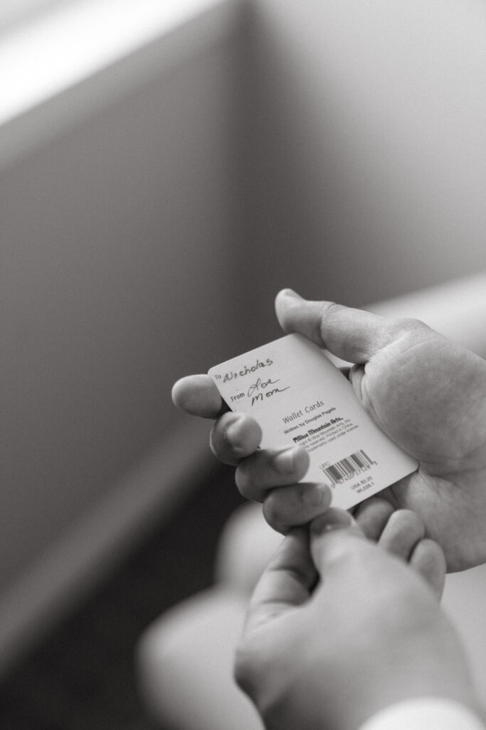 Close-up of the groom holding a small gift card that reads “To Nicholas, from Mom, Love Mom” as a sentimental moment before the ceremony.