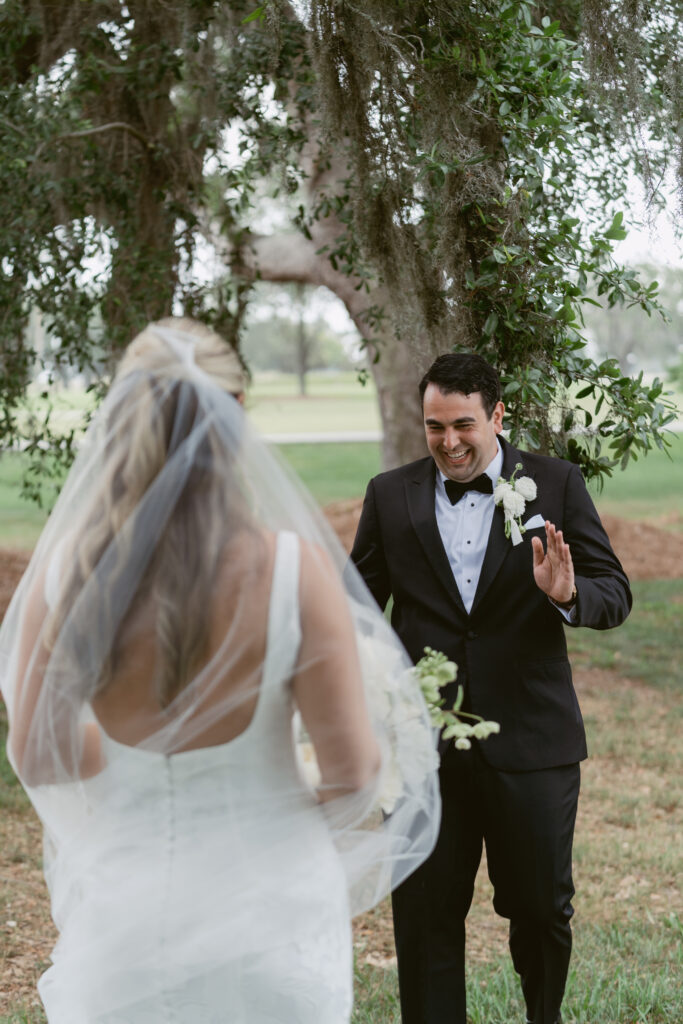 Groom smiling and raising his hand in excitement as he sees his bride for the first time during their emotional first look outdoors.