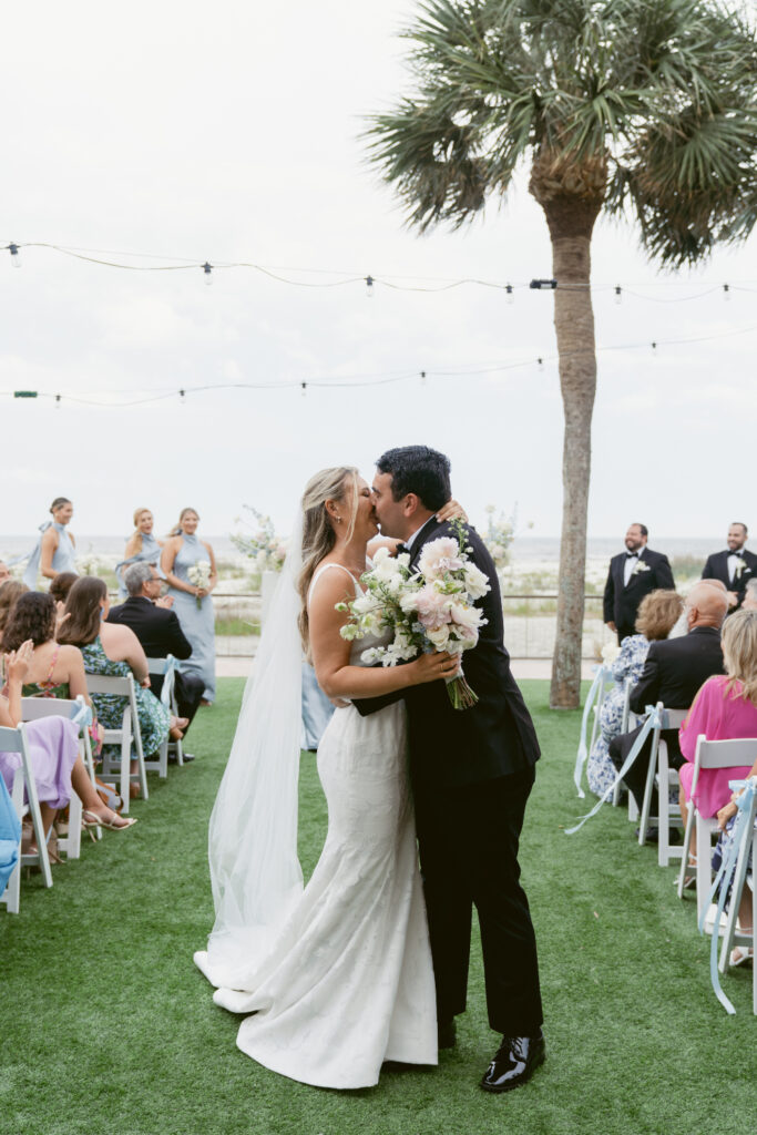Bride and groom share a kiss at the end of the aisle, holding a bouquet of soft pastel flowers with palm trees and guests in the background.