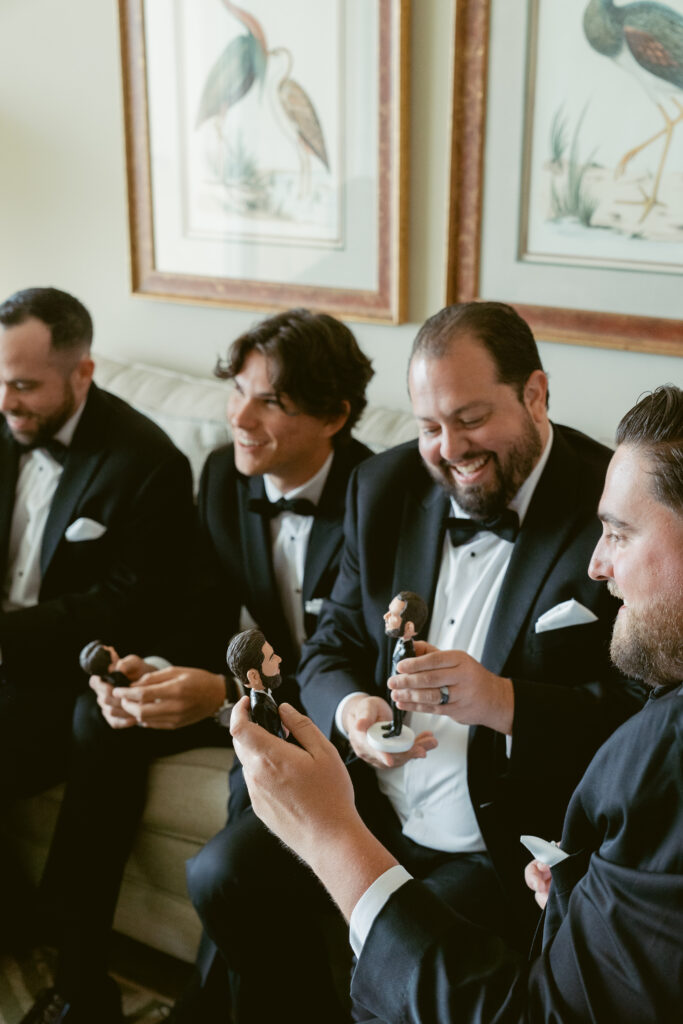 Groomsmen dressed in black tuxedos sit on a couch, smiling and laughing while holding personalized bobblehead figurines.